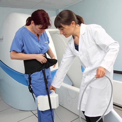 A practitioner scans the knee of a patient wearing a hip brace using an external machine, next to the upright open mri